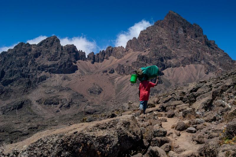 A porter carrying a load on his head up a rocky path below the jagged peaks of Mawenzi