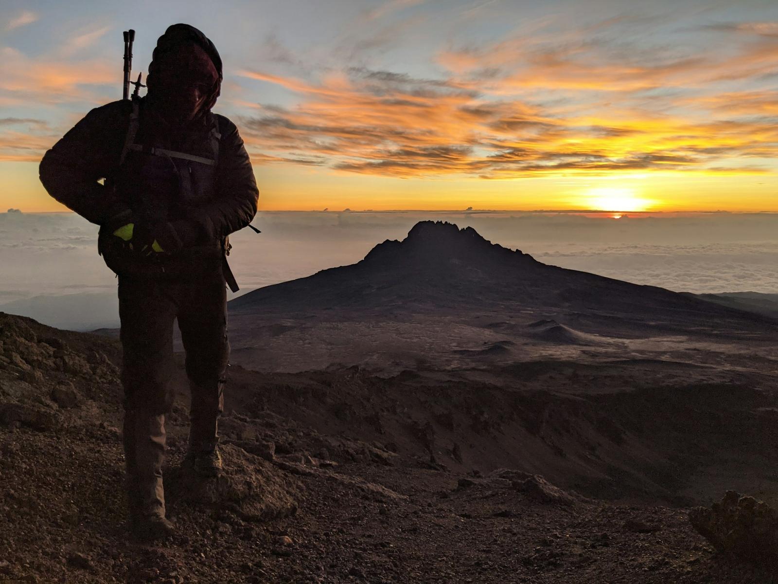 A climber silhouetted against the sunrise high on Kibo, with the jagged peak of Mawenzi rising above a sea of clouds